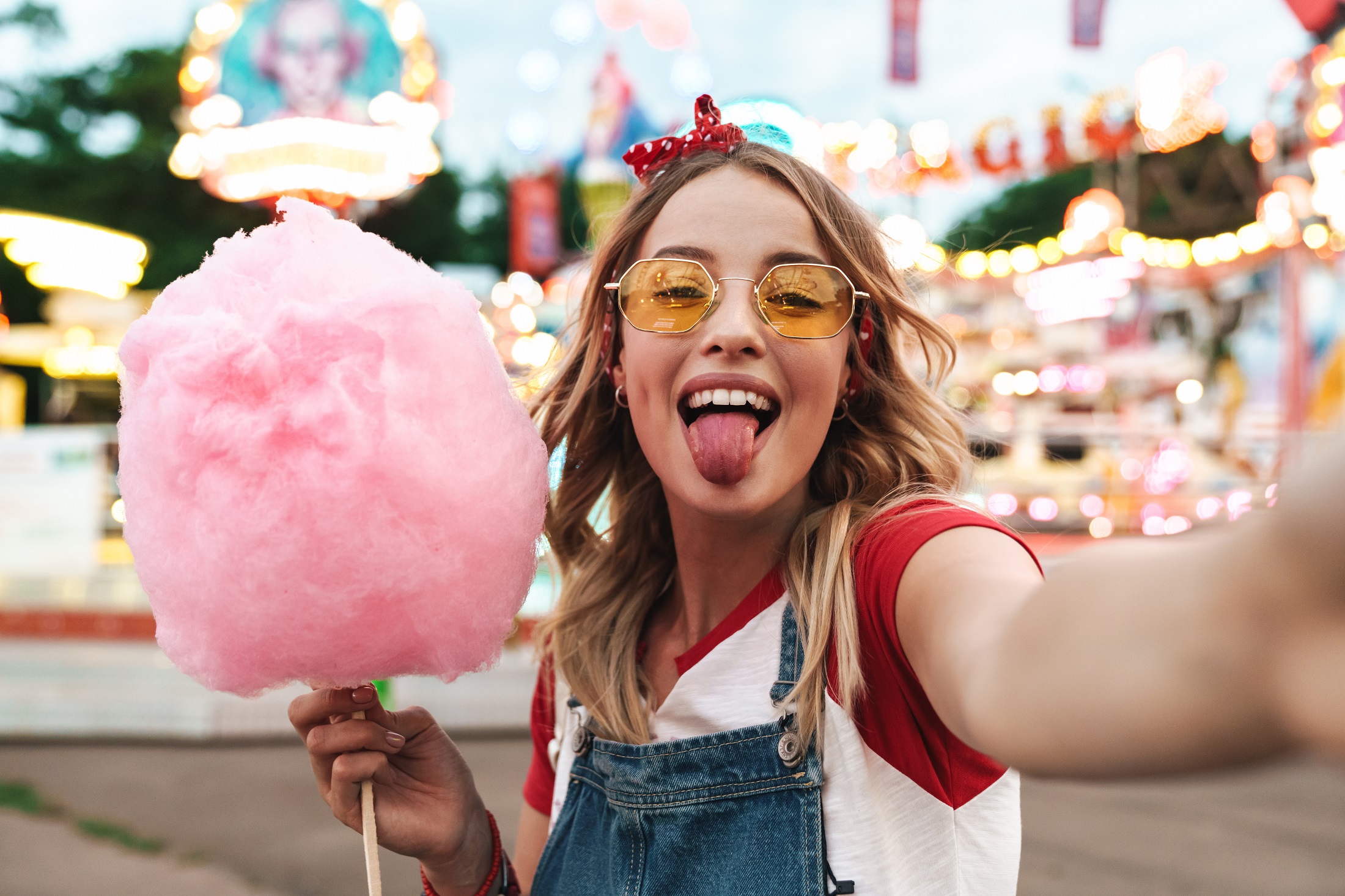 Image of positive blonde woman holding sweet cotton candy while taking selfie photo at amusement park